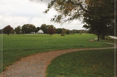 A peaceful landscape showing a winding path through a green field with scattered trees and a distant white house.